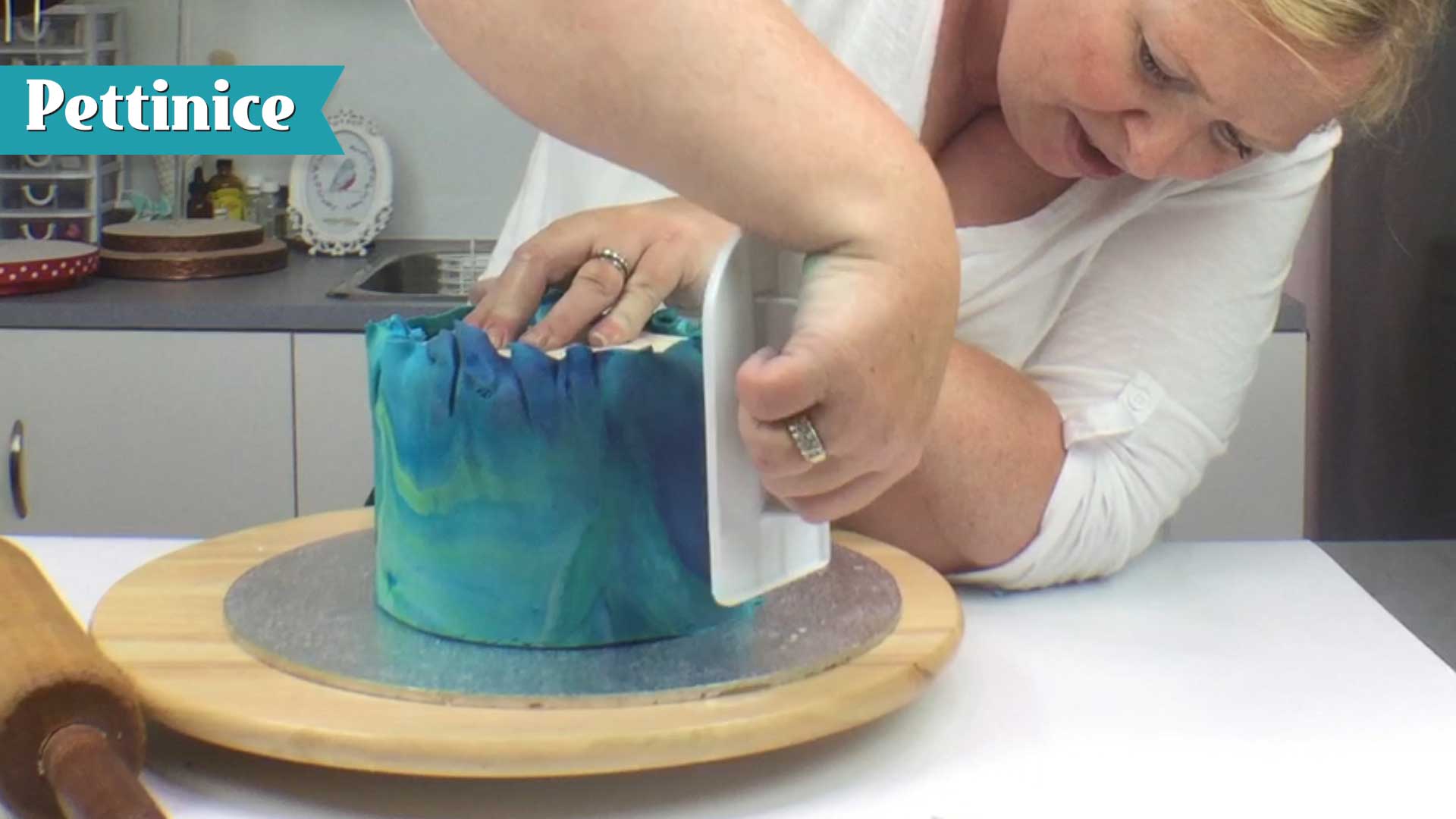 Use your cake smoother to push fondant to the board, working your way around the cake.  You will trim off the excess next.  We will demonstrative removing the folds shown here.  Because we used Sprink, it allows us more working time with our fondant.